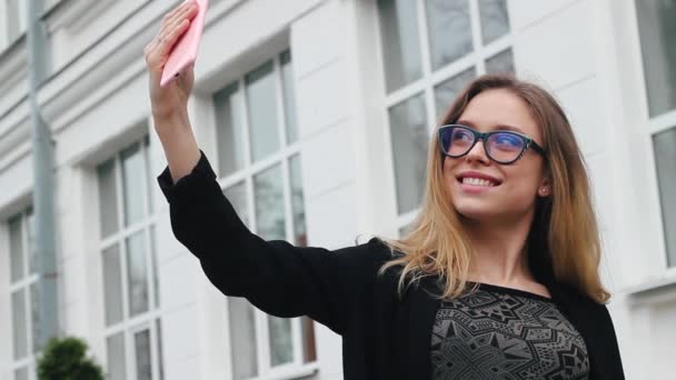 Sehr schönes und attraktives Mädchen mit Brille, das Selfie in der Nähe der Schule macht. Frau lächelt und fotografiert sich am Telefon. Sie ist glücklich — Stockvideo