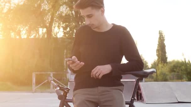 Handsome Guy Digite uma mensagem em pé perto de uma bicicleta ao pôr do sol . — Vídeo de Stock