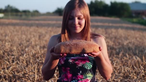 Mädchen, das in einem Weizenfeld steht und das Brot in seinen Händen hält. sie schnuppert und lächelt. Zeitlupe. — Stockvideo