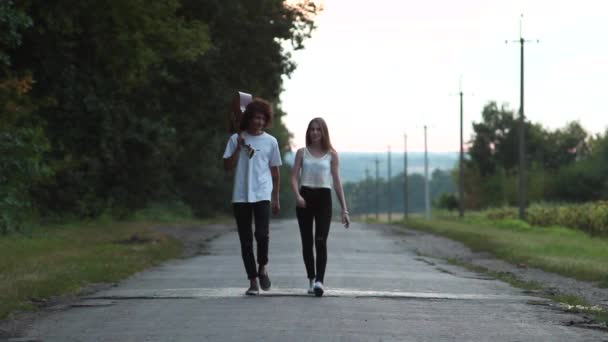 Pareja caminando por la carretera. Hombre sosteniendo una guitarra. Hipsters al atardecer . — Vídeos de Stock