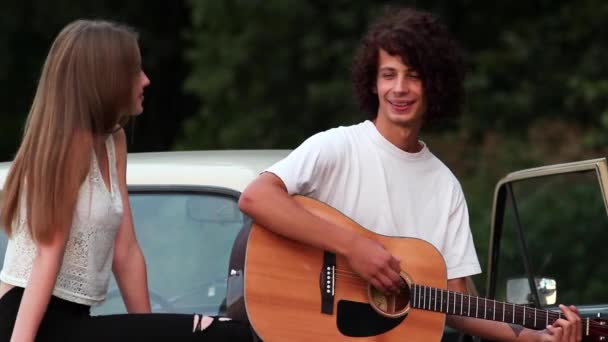 Den killen som spelar gitarr för en flicka sitter på Retro bilar. ett par mycket vackra. — Stockvideo