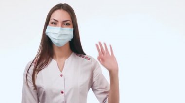 Güzel kadın doktor el sallıyor. Doktor yüzünde bir maske ile beyaz bir arka plan üzerinde duruyor.