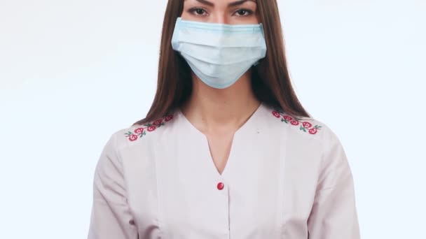 Mooie verpleegster in een ziekenhuis en kijkt naar de camera. Een meisje staat op een witte achtergrond met een masker op zijn gezicht. Ze is erg mooi. — Stockvideo