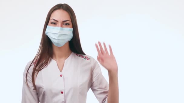 Красива жінка-лікар махає. Лікар стоїть на білому тлі з маскою на обличчі . — стокове відео