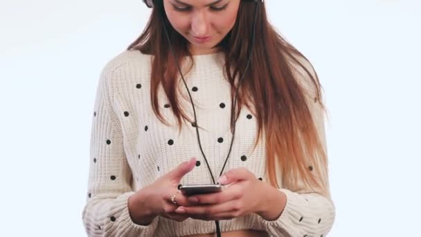 Hermosa chica escuchando música en los auriculares con el teléfono. La chica tiene un teléfono en la mano. Ella es muy atractiva. Fondo blanco . — Vídeo de stock