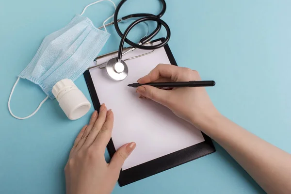 Top view of woman hands with pen over paper on folder isolated on blue background. Stethoscope, pills bottle and surgery mask with hands and paper on blue. Doctor fills out a form on paper for analyzes.