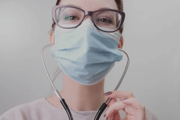 Close up portrait of young woman doctor cover her face wearing facial medical blue mask and glasses takes care of the examination. Doctor listens to the patient\'s lungs and bronchi with a stethoscope