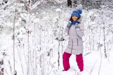 Kış şapkalı, eldivenli ve kar taneleriyle kaplı atkısı olan güzel, mutlu bir genç kadın. Kış ormanı manzarası geçmişi