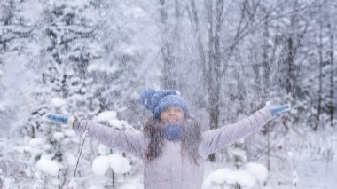 Kış şapkalı, eldivenli ve kar taneleriyle kaplı atkısı olan güzel, mutlu bir genç kadın. Kış ormanı manzarası.