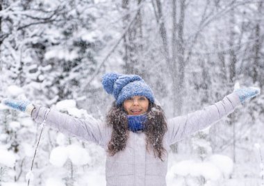 Kış şapkalı, eldivenli ve kar taneleriyle kaplı atkısı olan güzel, mutlu bir genç kadın. Kış ormanı manzarası.