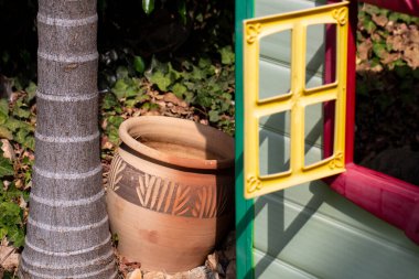 Büyük kahverengi kil bir sürahi bir palmiye ağacının yanında durur ve küçük sevimli bir evi olan çocuklar.