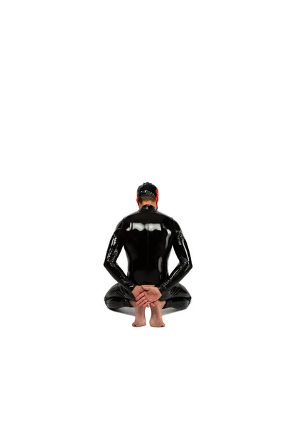 Fashion BDSM lateks catsuit içinde dizlerinin arkasında duran bir kölenin arka plan portresi izole edilmiş beyaz arka planda kırmızı maske takıyor. Süslü kauçuk fetişist giysiler giymiş bir adam.