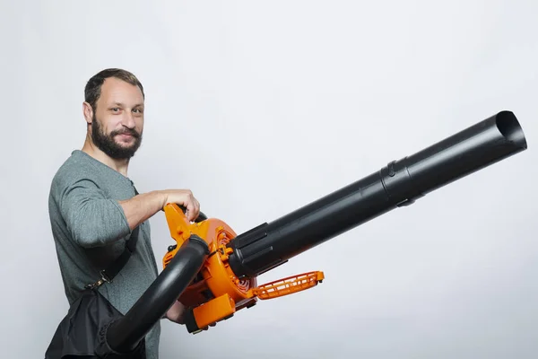 Yakışıklı sakallı adam elinde bahçe üfleyicisi ve gri arka planı olan bir çanta tutuyor. Elinde yaprak üfleyicisi olan bir işçi. Yaprak üfleyici ve toz filtresi. El bilgisayarı yaprakları savuruyor. Araçlar