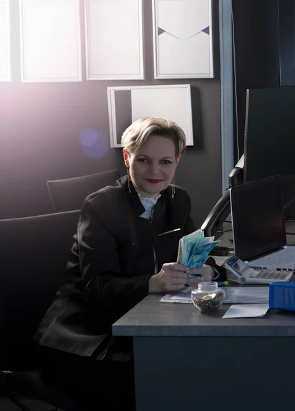 beautiful blonde woman in business suit, smiling at desk behind laptop computer and papers in home office. Business lady with New Israeli shekels and Dollars bills in hands, creative neon color.