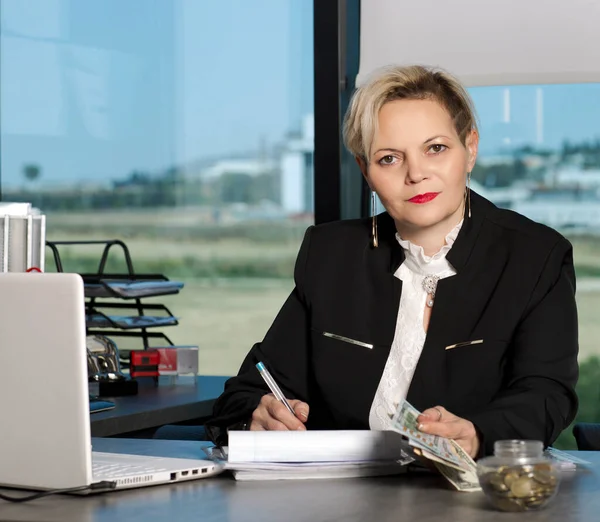 beautiful blonde woman in business suit, smiling at desk behind laptop computer and papers in office. Business lady with dollars banknotes in hand. Office worker writing with ballpoint pen in document