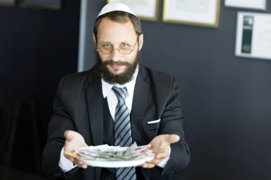 Bearded Jewish man in white Yarmulke (hat, Kippah) looking with cunning eyes. Businessman holding tray with money. Sly Jewish man smiling cunning looking at the camera. Selective focus on the eyes clipart