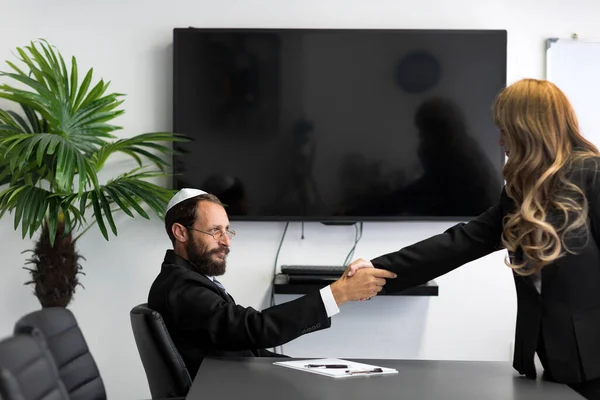 Shake Hands Over Desk. Jewish man in a yarmulke and glasses working at the office. Jew man in shirt, business suit and national hat Kippah shakes hands with a businesswoman, selective focus.