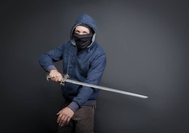 American Brutal man in a blue hoodie and black face mask with a Japanese sword. Portrait of a courageous warrior in jeans and a hood standing and holding sword in hand isolated on gray background
