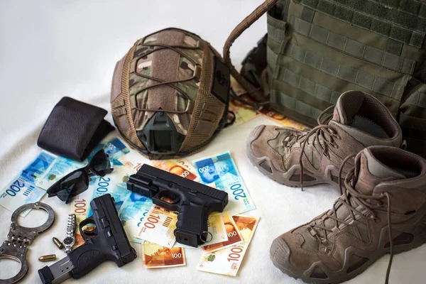 Military uniform and equipment, ammunition. Body armor, guns, bulletproof helmet, handcuffs, sunglasses and military boots with banknotes of Israeli New Shekels. Weapons for airsoft and urban protests