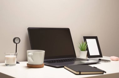 Beyaz bir masadaki dizüstü bilgisayarın resmi bir kupa bir saat küçük bir çalı bir defter bir telefon çerçevesi ve bir kulaklık.