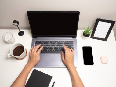Bir kadının elleri beyaz bir masanın üzerinde, klavyede dizüstü bilgisayarla bir cep telefonunun yanında siyah bir defter bir fincan kahve bir saat beyaz bir mum küçük bir çalılık ve boş bir fotoğraf çerçevesi.