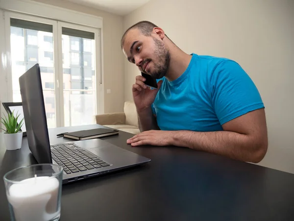Oturma odasından çalışan genç bir adam siyah bir masada dizüstü bilgisayarında beyaz bir mumla telefonu ve siyah defterli bir kredi kartıyla çalışıyor.