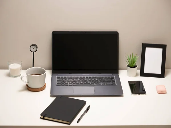 笔记本电脑在白桌子上的图片 上面有一个杯子 一个钟 一个小灌木丛 一个笔记本 一个电话 一个相框和一个耳机 — 图库照片
