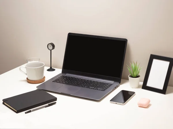 Beyaz bir masadaki dizüstü bilgisayarın resmi bir kupa bir saat küçük bir çalı bir defter bir telefon çerçevesi ve bir kulaklık.