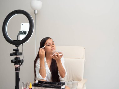 Beyaz bir masa ve halısı olan bir sandalyede farklı kadın ürünleriyle makyaj blogu yazan genç Latin kız.