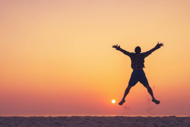 Günbatımı gökyüzü ve ada arka planıyla tropikal sahilde atlayan bir adam. Özgürlük iyi hissettirir ve seyahat macerası konsepti. Klasik ton filtre efekti renk biçimi.