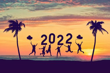 Günbatımı gökyüzünde 2022 numaraya atlayan siluet arkadaşları tropik plajda palmiye ağacının soyut arka planına sahipler. Mutlu yıllar ve bayram kutlamaları. Klasik ton rengi.