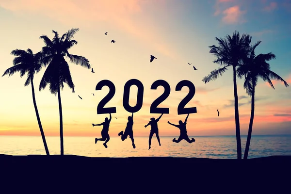 Silhouette friends jumping and holding number 2022 on sunset sky with palm tree birds flying background at tropical beach. Happy new year and holiday celebration concept. Vintage tone color style.