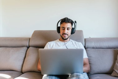 Kulaklıklı sakallı adam evdeki kanepede oturan dizüstü bilgisayarını kullanıyor. Çocuk online ağ oyunu oynuyor. İş yerindeki sanal bir buluşmayla bağlantılı koyu saçlı bir adam..