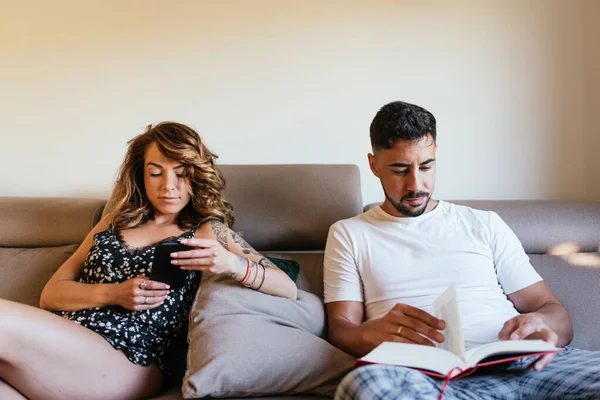 Happy couple sitting on the sofa at home with book and mobile phone. Man reading a book sitting at home. Woman sitting in the living room connected online with her smartphone. Family life