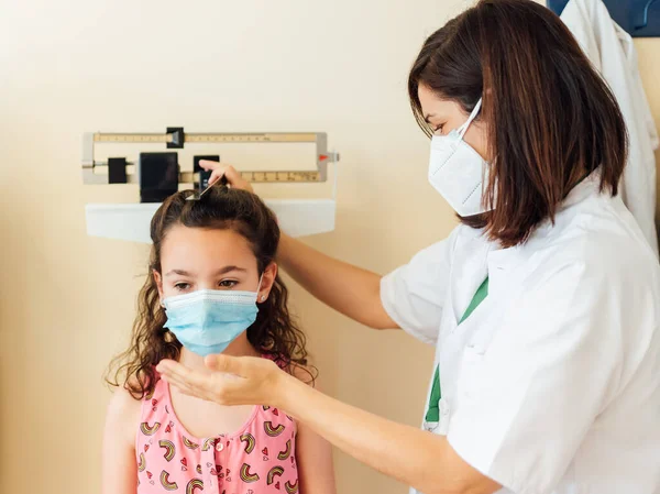 Çocuk doktoru klinikteki çocuğu ölçer ve tartır. İkisi de kovid-19 virüsünden korunmak için maske takıyor. Genç kız nasıl büyüdüğünü görmek için hastanede muayene ediliyor..