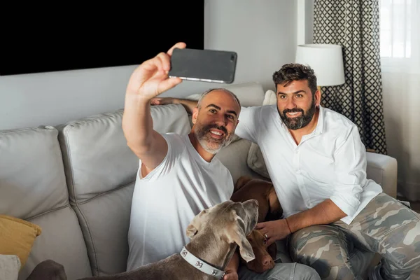 Aynı cinsten iki erkek oturma odalarında selfie çekiyor. Mutlu, gülümseyen, beyaz tenli adamlar evlerinde köpekleriyle eğleniyorlar. lgtbi kavramı