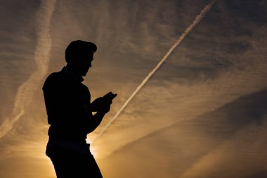 Şehirde gün batımında cep telefonu olan genç bir çocuğun silueti.
