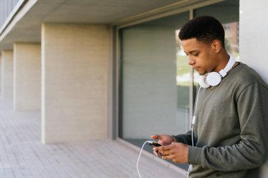 Afro çocuk sokakta kulaklıklarıyla müzik dinlerken cep telefonuna bakıyor..