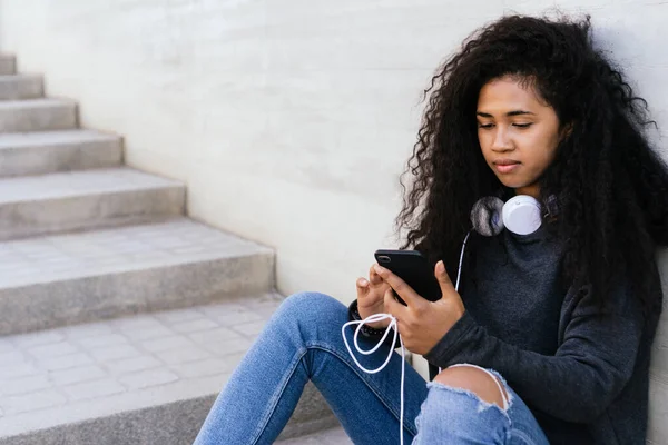Güzel kıvırcık saçlı afro kız sokakta oturmuş kulaklıklı cep telefonu kullanıyor.