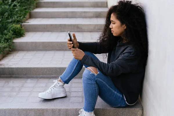 Menina afro sentado navegando na Internet com seu telefone celular — Fotografia de Stock