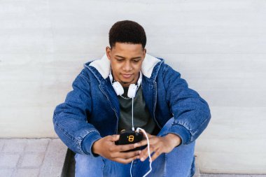 Sokakta cep telefonu ve kulaklığını kullanan Latin bir adamın portresi. Gri bir duvarda cep telefonu kullanan genç afro bir çocuk. Kopyalama alanı olan yatay fotoğraf 