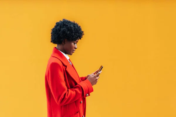 Portrait of black woman with wavy hair holding in hands mobile chat device isolated on bright yellow background. Afro girl walking with cell phone on plain background. horizontal photo, copy space.