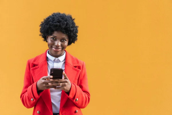 Happy young African American girl with afro hair and red coat standing on yellow background holding smart phone looking at camera. Black woman using smart phone app. afro girl connected to internet.