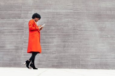 Kırmızı ceketli zarif afro kadın gri arka planda tablet giyiyor. Siyah kız internete bağlı taşınabilir cihaz kullanıyor. İletişim konsepti. Boşluğu kopyala.