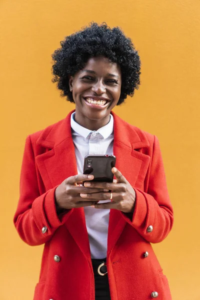 Sarı arka planda duran afro saçlı ve kırmızı ceketli mutlu Afro-Amerikalı kız akıllı bir telefon tutuyor.. 