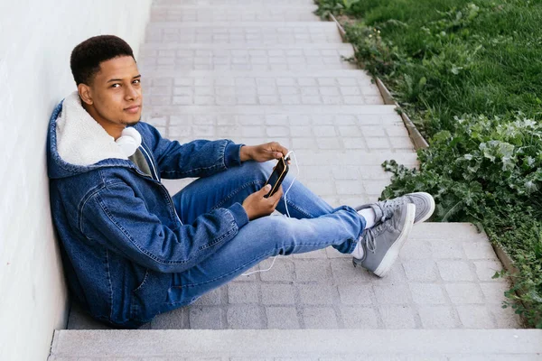 Afro menino na rua usando seu telefone celular com fones de ouvido. — Fotografia de Stock