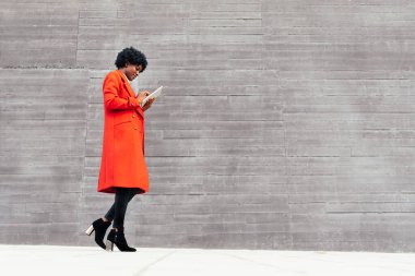 Kırmızı ceketli zarif afro kadın gri arka planda tablet giyiyor.