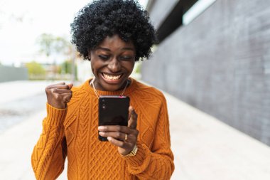Mutlu ve duygusal Afrikalı Amerikalı bir kadının görüntüsü. Arka planda izole edilmiş bir şekilde cep telefonuyla zafer jesti yapıyor..