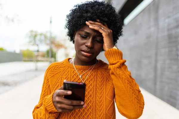 Endişeli Afrikalı Amerikalı kadın sokakta cep telefonunu kontrol ediyor. Kıvırcık saçlı esmer kız huzursuz ve gergin hareketleriyle.