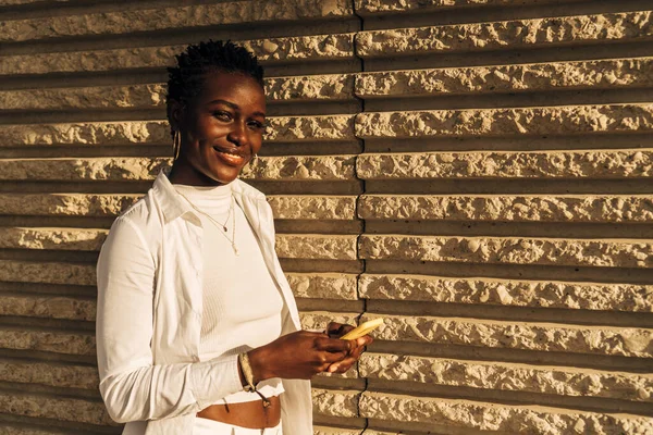 Mulher afro-americana sorrindo e olhando para a câmera enquanto segurava um telefone celular. — Fotografia de Stock